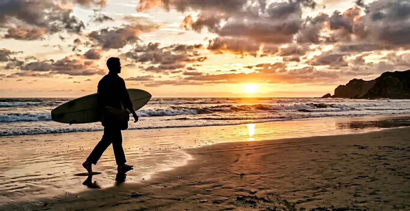 Business professional carrying surfboard walking towards ocean at sunset with laptop bag balanced on shoulder